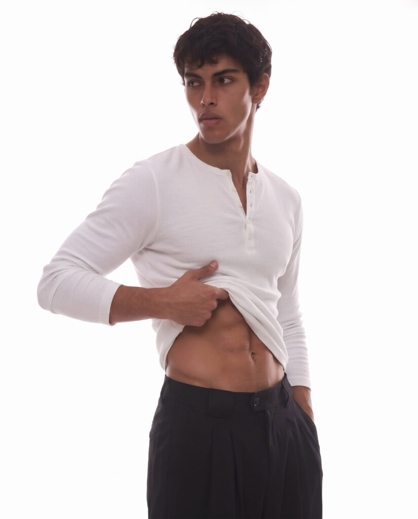 Male model in white long-sleeve shirt lifting to show abs, black pants, minimal background.