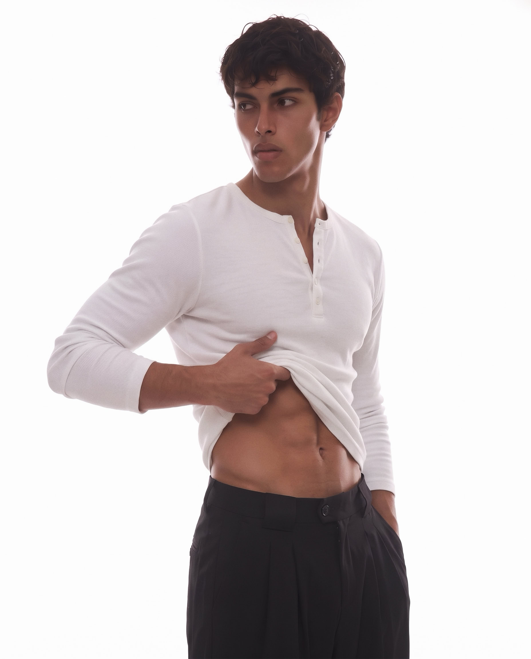 Male model in white long-sleeve shirt lifting to show abs, black pants, minimal background.